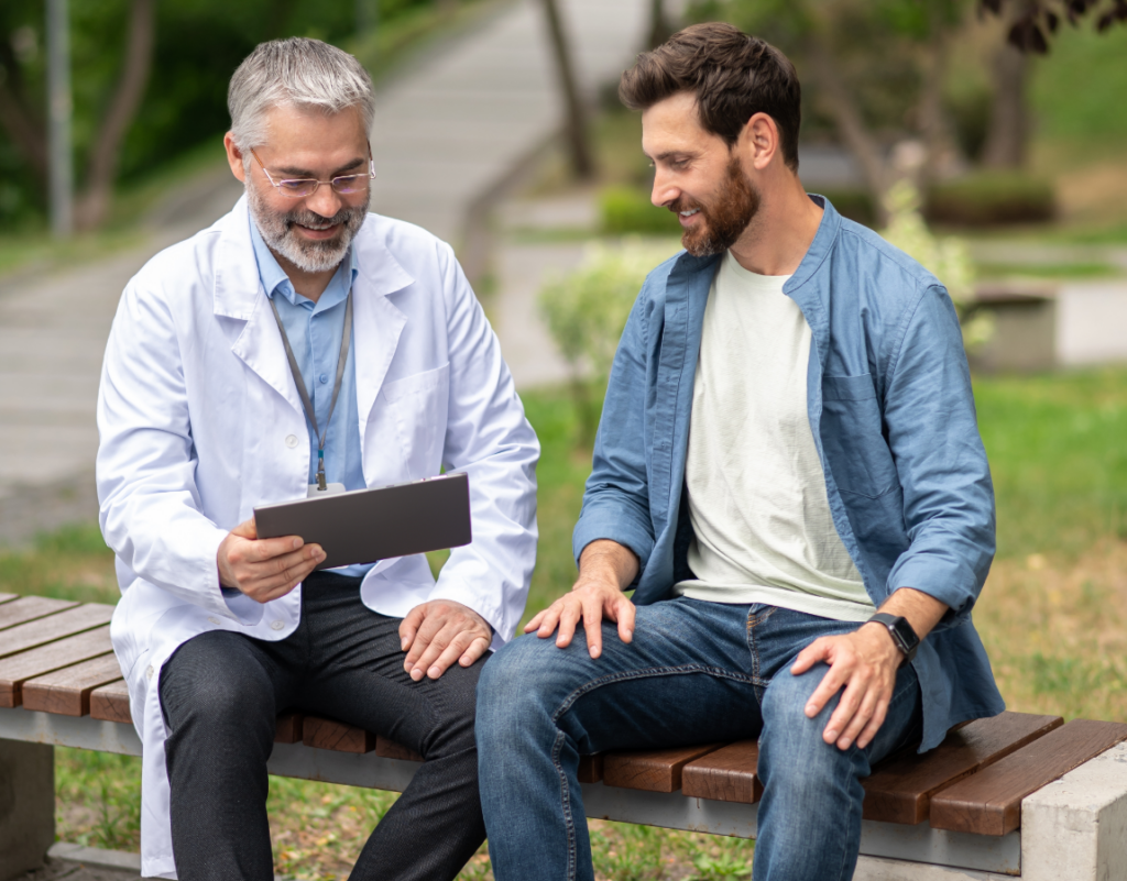 A doctor in a white coat explains hyperbaric oxygen therapy on a tablet to a smiling man beside him on a park bench; both appear engaged and relaxed in the outdoor setting.