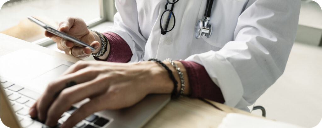 A person wearing a white lab coat and stethoscope uses a smartphone while typing on a laptop, suggesting they are a healthcare professional multitasking at a desk, possibly coordinating a clinical trial for hyperbaric oxygen therapy.