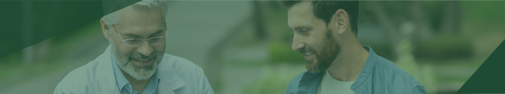 A smiling doctor in a white coat and glasses talks with a bearded man outdoors. Both appear engaged in a pleasant conversation. The background is blurred greenery.